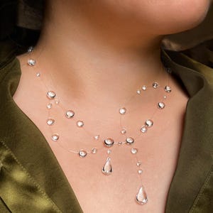 May include: A delicate necklace featuring clear, round beads and teardrop-shaped pendants. The beads are strung on a nearly invisible cord, creating a floating effect. The necklace is worn against a green background.