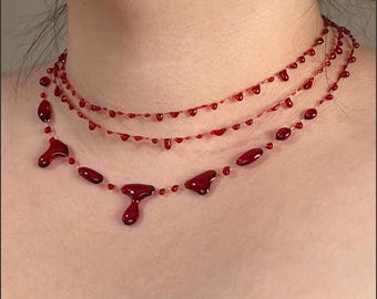 May include: A three-strand necklace featuring red, teardrop-shaped beads. The beads are strung on a thin, clear cord, creating a delicate and eye-catching design. The necklace is worn around the neck.
