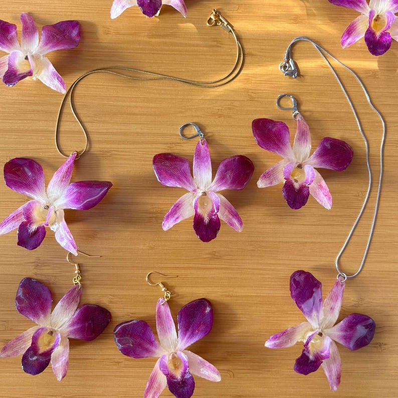May include: A set of jewelry featuring purple and white orchid flower charms. The set includes a necklace, earrings, and a pendant. The jewelry is displayed on a wooden surface.