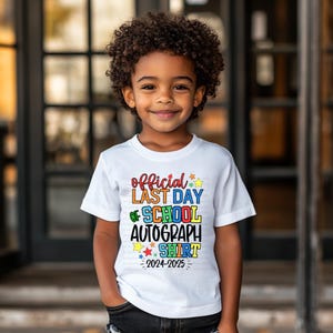 May include: A white t-shirt with the text "official last day of school autograph shirt 2024-2025" in colourful letters and stars. A young child is wearing the shirt.