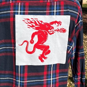 May include: A blue and red plaid flannel shirt with a white patch featuring a red fire demon graphic.