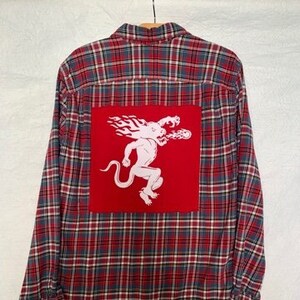 May include: Red, blue, and white plaid flannel shirt with a white graphic on the back. The graphic is a white creature on a red square. The shirt is hanging on a wooden hanger.