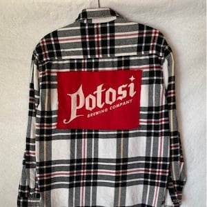 May include: A white and black plaid flannel shirt with a red patch on the back that says "Potosi Brewing Company" in white lettering.