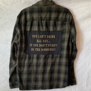 May include: A green and black plaid flannel shirt with a patch on the back that reads "You can't drink all day... If you don't start in the morning!"