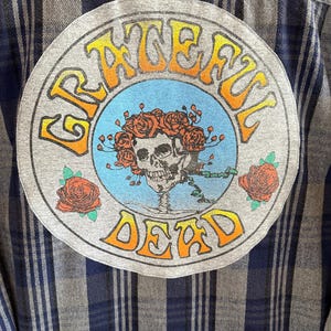 May include: A gray and blue plaid flannel shirt with a circular patch on the back. The patch features the words "Grateful Dead" in yellow and orange lettering with a skull and roses design inside.