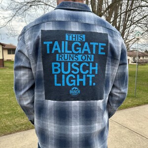 May include: A blue and white plaid flannel shirt with a black patch on the back that says "This tailgate runs on Busch Light." The patch has a small Busch Light logo in the bottom right corner.