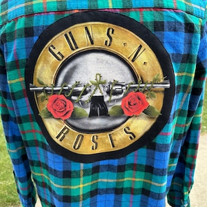 May include: A blue, green, and black plaid flannel shirt with a Guns N' Roses logo on the back. The logo features a gold-colored emblem with a gun and roses design, and the band's name in a circular arrangement.