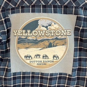 May include: A blue and white plaid flannel shirt with a patch on the front. The patch is a light blue circle with a brown mountain scene and the text "Yellowstone" in gold letters. The text "Dutton Ranch Montana" is printed below the circle.