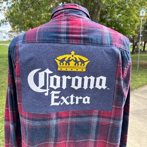May include: A red and blue plaid flannel shirt with a blue patch on the back featuring a white Corona Extra logo with a gold crown.