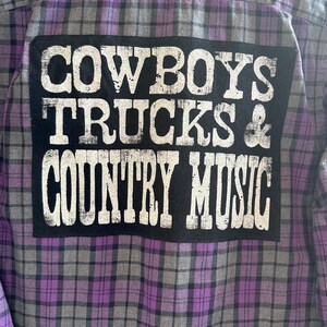 May include: A purple and gray plaid flannel shirt with a black patch on the back that says "COWBOYS TRUCKS & COUNTRY MUSIC" in white letters.