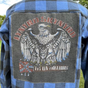 May include: Blue and black plaid flannel shirt with a large Lynyrd Skynyrd patch on the back. The patch features an eagle with a Confederate flag in the bottom left corner and the text "Lynyrd Skynyrd" and "One More From The Bird" in a circle around the eagle.