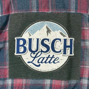 May include: A red, blue, and white plaid flannel shirt with a dark gray patch. The patch features a circular design with the words "BUSCH Latte" in blue and white, with a mountain scene. The text "Anheuser-Busch St. Louis, MO" is also visible.