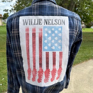 May include: A blue and white plaid flannel shirt with a white patch on the back. The patch features a red, white, and blue American flag design with guitar necks and pick-ups in place of the stripes. The text "Willie Nelson" is printed above the flag.