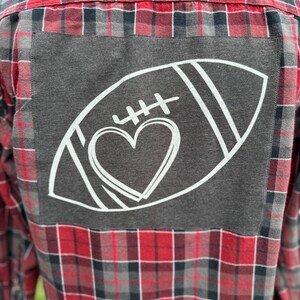 May include: A red and black plaid flannel shirt with a gray patch on the back. The patch features a white outline of a football with a heart inside.