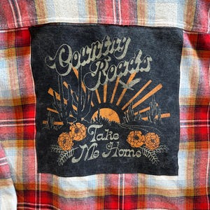 May include: A red and black plaid flannel shirt with a black patch sewn on the front. The patch features a sunset graphic with the text "Country Roads Take Me Home".
