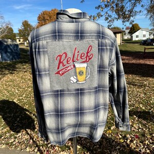 May include: A gray and navy blue plaid flannel shirt with a gray patch on the back. The patch features the words "Relief Pitcher" in red script, a baseball, and a beer mug.