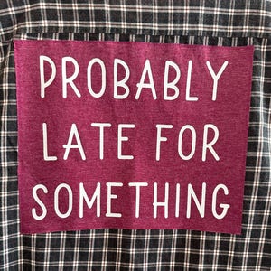 May include: A burgundy plaid shirt with a pink patch on the back that says "PROBABLY LATE FOR SOMETHING" in white text.