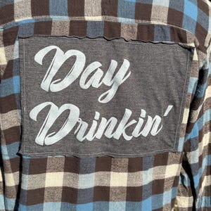 May include: A brown, blue, and white plaid flannel shirt with a gray patch sewn on the back. The patch has the words "Day Drinkin'" written in white.