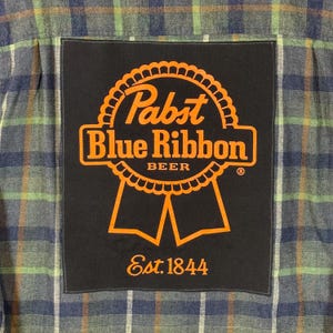 May include: A plaid button-up shirt with a black patch featuring the Pabst Blue Ribbon Beer logo in orange. The patch includes the text "Est. 1844". The shirt has a blue, green, and white plaid pattern.