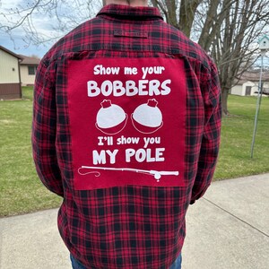 May include: A red and black plaid flannel shirt with a red patch on the back. The patch has white text that says "Show me your Bobbers" and a graphic of two fishing bobbers. The text below the bobbers says "I'll show you my pole" and a graphic of a fishing rod.