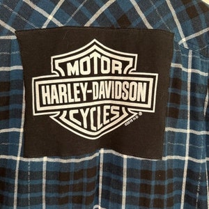 May include: Blue and white plaid flannel shirt with a black patch featuring the Harley-Davidson logo. The logo is white with the words "MOTOR", "HARLEY-DAVIDSON", and "CYCLES".