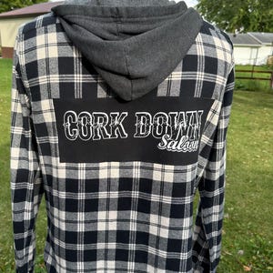 May include: Black and white plaid flannel shirt with a gray hooded sweatshirt attached. The back of the shirt has a black patch with the text "Cork Down Saloon" in white.