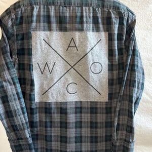 May include: A gray and black plaid flannel shirt with a gray patch on the back that says "W A O C" in black letters.