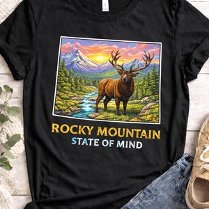 Może przedstawiać: Czarny t-shirt z ilustracją górskiego krajobrazu, jelenia, rzeki i zachodu słońca. Tekst "ROCKY MOUNTAIN STATE OF MIND" jest wydrukowany poniżej. Swobodny t-shirt.