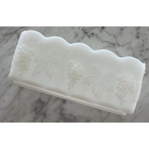 May include: A rectangular white milk glass planter featuring a scalloped top edge and an embossed pattern of grape clusters and vine leaves wrapping around the exterior sides. The vessel has a smooth, opaque finish and a flat base.