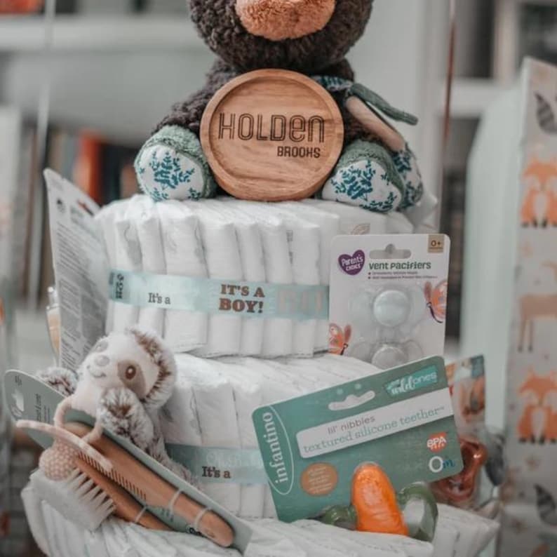 May include: A multi-tiered diaper cake decorated with a brown teddy bear wearing a green and blue patterned outfit, a wooden sign that reads "Holden Brooks", and blue and white ribbon. The cake includes a variety of baby items, such as pacifiers, teethers, and a wooden comb.