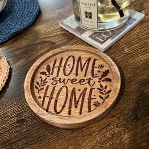 May include: A round wooden coaster with the text "Home sweet home" engraved on it. The coaster has a brown wood finish and features a decorative design of leaves and snowflakes.