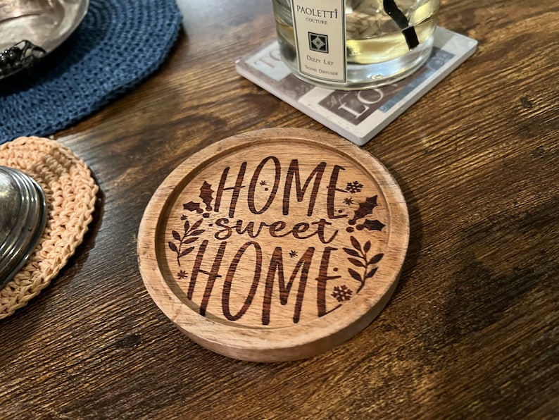 May include: A round wooden coaster with the text "Home sweet home" engraved on it. The coaster has a brown wood finish and features a decorative design of leaves and snowflakes.
