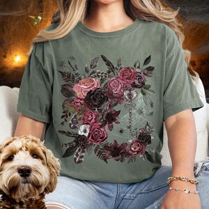 May include: A green t-shirt with a floral design featuring dark pink, red, and purple flowers, leaves, and feathers. A silver key and spider web are also included in the design.