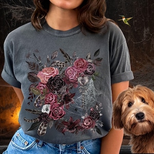 May include: A dark gray t-shirt with a floral design featuring dark red, pink, and purple roses, leaves, and feathers. A key and spiderwebs are also included in the design.