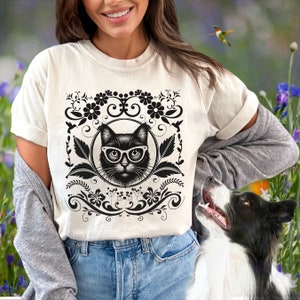 May include: A white t-shirt with a black and white graphic of a cat wearing glasses. The graphic is surrounded by a floral design.