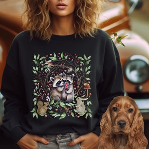 May include: Black sweatshirt with a watercolor illustration of two raccoons holding red roses, surrounded by green leaves, pink hearts, and a squirrel. The design is set against a dark background.