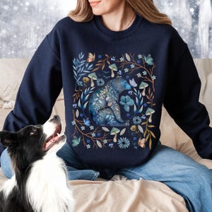 May include: A navy blue sweatshirt with a watercolor design of a bear surrounded by flowers, mushrooms, and butterflies.