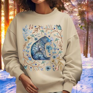 May include: A beige sweatshirt with a blue bear surrounded by flowers, mushrooms, and butterflies. The bear is sitting on a branch and has a floral pattern on its fur.
