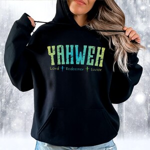May include: Black hooded sweatshirt with the word "YAHWEH" in distressed green and blue lettering. Below the word are the words "Lord Redeemer Savior" with cross symbols. The sweatshirt has a front pocket and a drawstring hood.