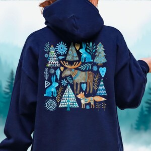 May include: Navy blue hoodie featuring a whimsical winter design. The back of the hoodie showcases a watercolor illustration of a moose, fox, rabbits, and trees in shades of blue, teal, and gold. The design includes snowflakes and heart shapes.