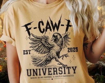 May include: Mustard yellow t-shirt with a black graphic of a raven with outstretched wings. The text "F-CAW-F" is above the raven, with "EST 2025" and "UNIVERSITY" below. The phrase "WHERE BEING NORMAL IS VASTLY OVERRATED" is at the bottom.