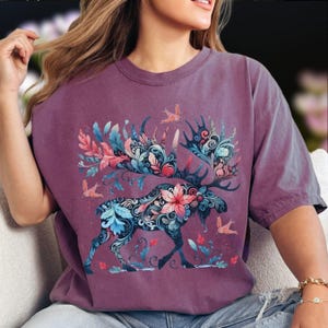 May include: A maroon t-shirt featuring a detailed illustration of a moose with floral antlers and body details. The design incorporates blue, pink, and teal floral elements, with birds in flight. The shirt has short sleeves and a relaxed fit.