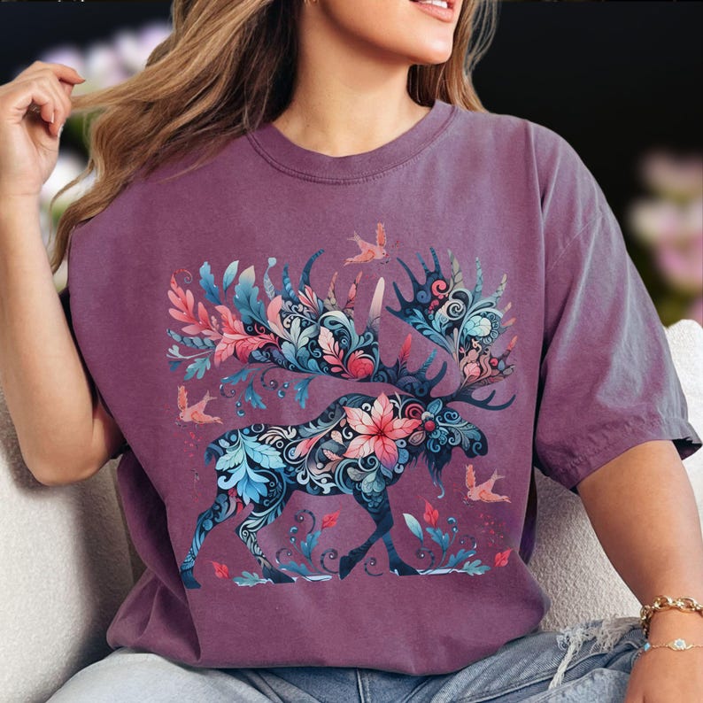 May include: A maroon t-shirt featuring a detailed illustration of a moose with floral antlers and body details. The design incorporates blue, pink, and teal floral elements, with birds in flight. The shirt has short sleeves and a relaxed fit.