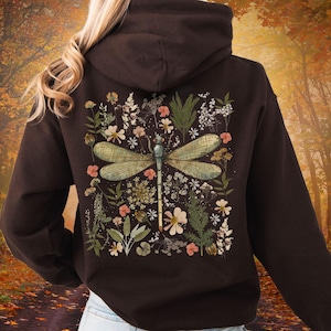 May include: A dark brown hoodie featuring a large dragonfly design on the back, surrounded by a floral arrangement of various flowers and greenery. The dragonfly has green wings and a blue body. The hoodie has a hood and a drawstring.
