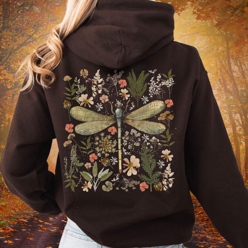 May include: A dark brown hoodie featuring a large dragonfly design on the back, surrounded by a floral arrangement of various flowers and greenery. The dragonfly has green wings and a blue body. The hoodie has a hood and a drawstring.