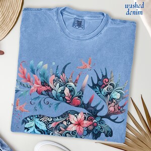 May include: Light blue t-shirt with a floral moose antler design. The antlers are filled with pink, teal, and blue floral patterns. The shirt has the words "washed denim" printed on it.