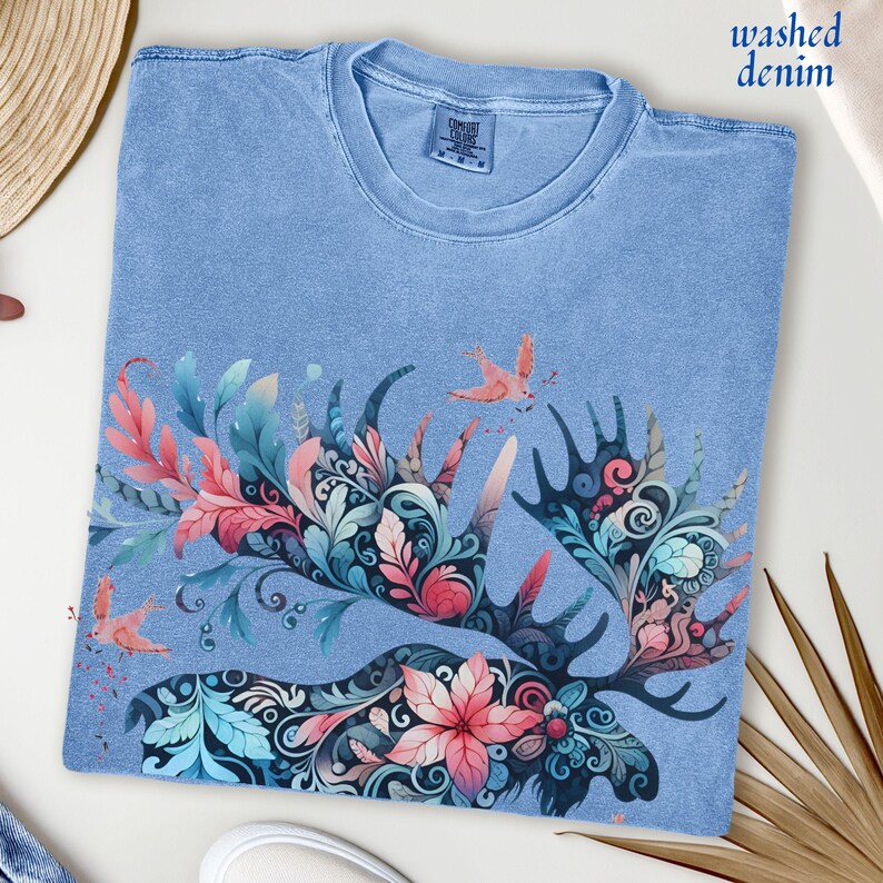 May include: Light blue t-shirt with a floral moose antler design. The antlers are filled with pink, teal, and blue floral patterns. The shirt has the words "washed denim" printed on it.