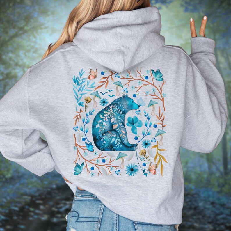 May include: A light gray hoodie featuring a watercolor design on the back. The design includes a blue bear surrounded by blue flowers, butterflies, and mushrooms. The hoodie has a hood and long sleeves.