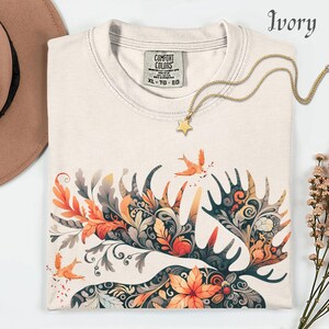 May include: Ivory-colored t-shirt with a detailed moose antler design in orange, gray, and teal. The shirt features a gold star pendant necklace and a Comfort Colors tag. The design includes birds and floral elements.