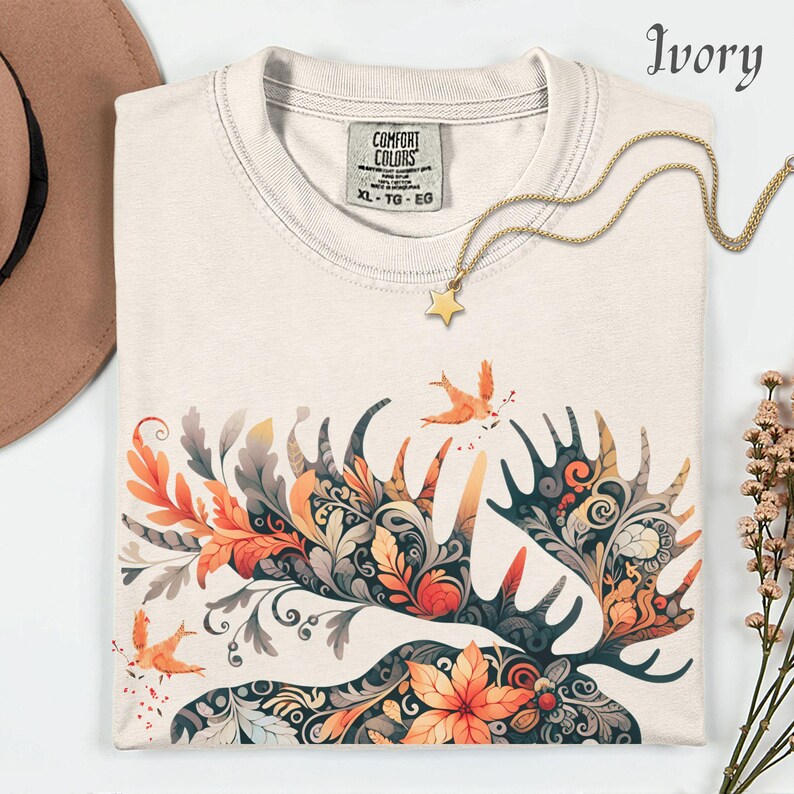 May include: Ivory-colored t-shirt with a detailed moose antler design in orange, gray, and teal. The shirt features a gold star pendant necklace and a Comfort Colors tag. The design includes birds and floral elements.